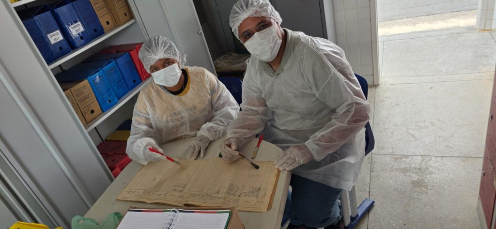 Fotografia de duas pessoas usando aventais, toucas e luvas descartáveis sentadas em uma sala com armários e prateleiras ao fundo. Elas manuseiam documentos antigos sobre uma mesa, indicando trabalho de análise ou preservação de arquivos.
