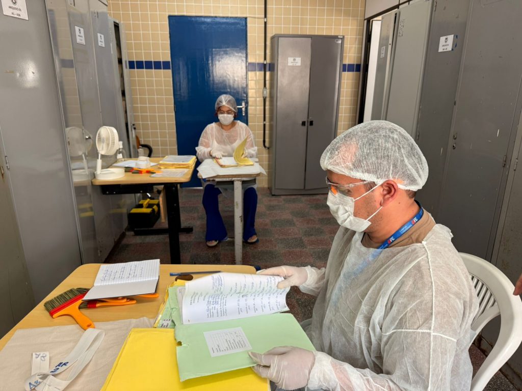 Fotografia colorida de ambiente de sala com arquivos da escola, com duas pessoas usando toucas, máscaras e aventais descartáveis, manuseando documentos e cadernos. Ambiente possui armários metálicos cinza, paredes com azulejos bege e azul, e mesas com materiais organizados.