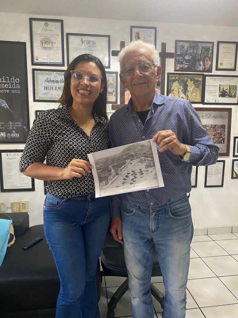 Fotografia de Renata e Onildo Almeida em ambiente interno segurando uma fotografia em preto e branco de uma paisagem com árvores e construções. Parede ao fundo exibe vários quadros com certificados, fotos e textos, destacando um crucifixo centralizado.