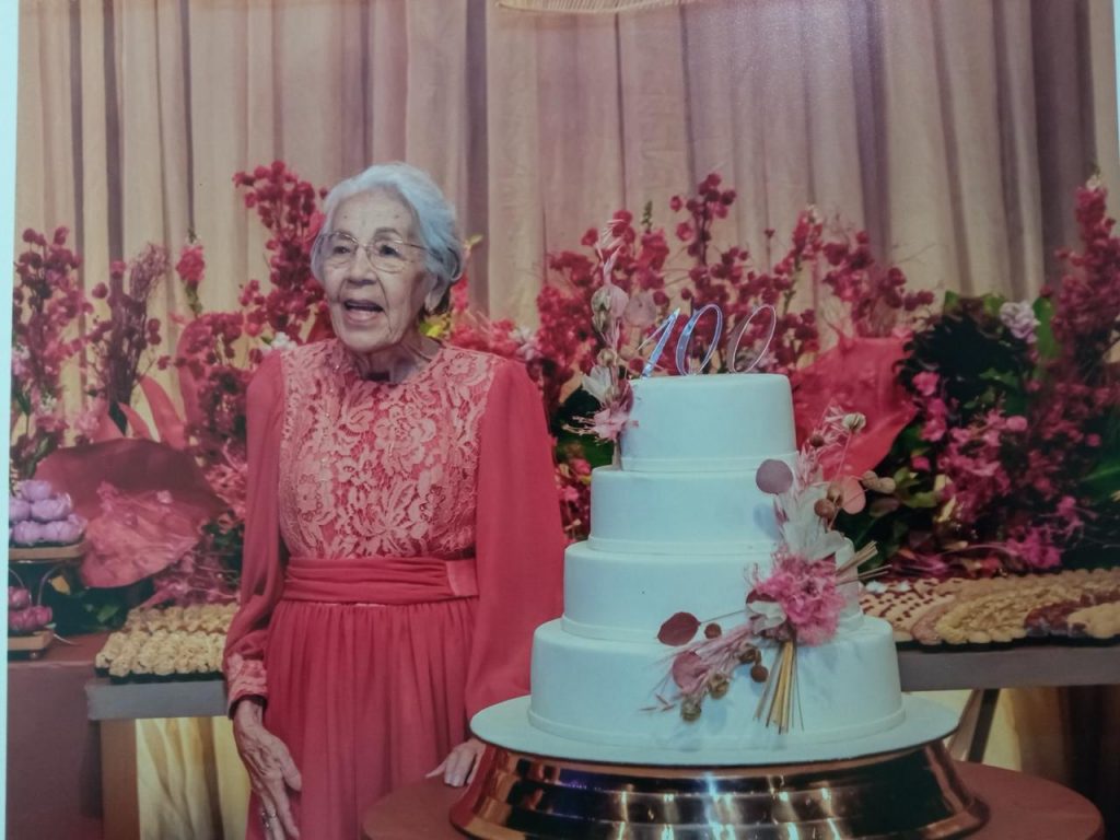 Fotografia de festa de aniversário de Marina Rocha vestindo vestido rosa posando ao lado de bolo branco de três andares decorado com flores e topo com número 100. Fundo apresenta decoração floral em tons de rosa e mesa com doces variados, indicando celebração centenária.