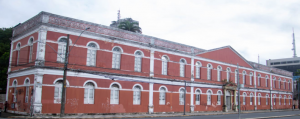 Fotografia colorida de prédio do ginásio pernambucano de dois andares com fachada vermelha e detalhes brancos, apresentando janelas arqueadas e ornamentos clássicos. Estrutura retangular ocupa esquina de rua, com elementos arquitetônicos que indicam uso institucional ou público.