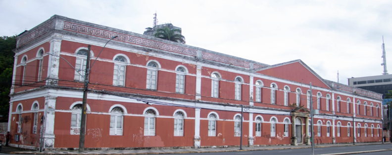 Fotografia colorida de prédio do ginásio pernambucano de dois andares com fachada vermelha e detalhes brancos, apresentando janelas arqueadas e ornamentos clássicos. Estrutura retangular ocupa esquina de rua, com elementos arquitetônicos que indicam uso institucional ou público.