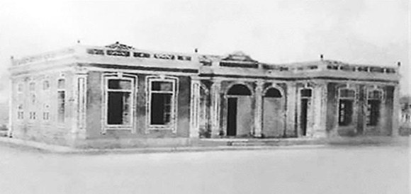 Fotografia em preto e branco do edifício do grupo escolar Joaquim Nabuco de arquitetura clássica com várias janelas retangulares e três arcos na entrada principal. Estrutura de um único andar com detalhes ornamentais no topo das paredes e fachada simétrica.